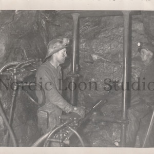May include: Black and white photograph of two miners in a tunnel. They wear helmets with lamps and work with machinery. The image is a vintage photo, likely from the early to mid-20th century.