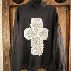 May include: A dark grey sweatshirt with a high neckline. The sweatshirt features a large, white, cross-shaped appliqué with a floral pattern in the centre. The cross is trimmed with a scalloped edge. The sweatshirt is hanging on a black hanger.