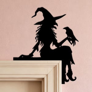 May include: A black silhouette wall decoration featuring a witch wearing a pointed hat and a long dress, sitting on a ledge. The witch is holding a raven perched on one hand. The design is flat and solid black, intended for placement on a corner.