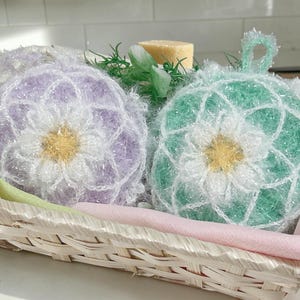 以下が含まれることがあります： 柳のバスケットには、手作りの花の形をした2つのスクラビーが入っています。1つはラベンダー色、もう1つはミントグリーン色で、白と黄色の中心があります。石鹸と緑の植物が背景にあります。スクラビーには吊り下げ用のループが付いています。