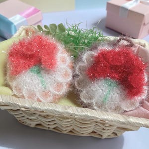 以下が含まれることがあります： 赤と白の花びらと緑色の中心部を持つ、手作りのふわふわした花型のアイテムが2つ、柳のバスケットに入っています。 バスケットは黄色の布で裏打ちされ、緑の小枝が入っています。 ギフトボックスが背景にあります。