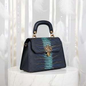 May include: A navy blue handbag with a textured crocodile pattern and a teal center stripe. The bag features a gold-colored clasp and handle attachments, and a black handle. The bag is sitting on a white marble surface.