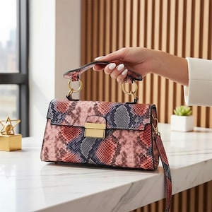 May include: A pink, red, and blue snakeskin-patterned handbag with a gold clasp and handle rings. The bag has a top handle and a detachable shoulder strap. The bag is sitting on a white marble surface.