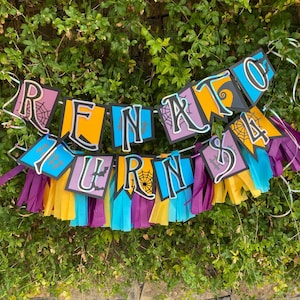 Puede incluir: Una colorida pancarta con el texto "RENATO TURNS 4" en letras negras, naranjas, moradas y azules. La pancarta está decorada con telarañas y borlas en morado, amarillo y azul. La pancarta se muestra sobre un fondo de hojas verdes.