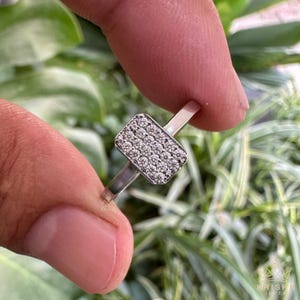 May include: A silver ring with a rectangular face covered in small, sparkling stones. The ring is held between two fingers against a backdrop of green foliage. The ring's band is thin and polished.