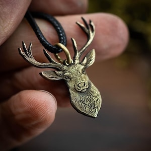 Könnte beinhalten: Halskette mit einem bronzefarbenen Hirschkopf-Anhänger. Der Anhänger hat detaillierte Geweihe und Gesichtszüge und hängt an einer schwarzen Kordel. Der Hintergrund ist unscharf, was das Design hervorhebt.