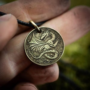 Könnte beinhalten: Runder Anhänger aus Bronze mit einem detaillierten Drachendesign. Der Anhänger hängt an einem schwarzen Band. Der Drache mit Flügeln und gewundenem Körper erzeugt ein Fantasy-Thema.