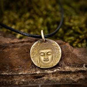 Puede incluir: Un collar con colgante dorado con un relieve detallado del rostro de Buda. El colgante redondo está suspendido de un cordón negro. El fondo es una vista borrosa de madera y musgo verde.