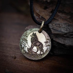 May include: A round, bronze-colored pendant necklace featuring a detailed engraving of a wolf standing on a rock, set against a backdrop of a full moon and trees. The pendant hangs from a black cord.