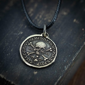Puede incluir: Collar con colgante plateado con un diseño de calavera y huesos cruzados. El colgante redondo tiene las palabras "MEMENTO MORI" alrededor del borde. El collar tiene un cordón negro.