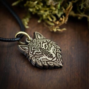 Op de afbeelding: Houten hangerketting met een gedetailleerd wolfskopontwerp. De donkerbruine hanger heeft ingewikkelde lichtgekleurde gravures en hangt aan een zwart koord met een goudkleurige sluiting. De achtergrond is een vage houten ondergrond.