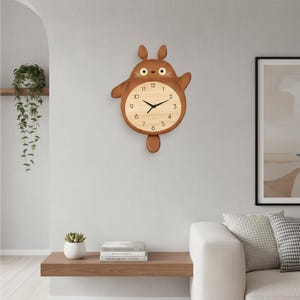 Könnte beinhalten: Eine Holz-Wanduhr in Form einer Zeichentrickfigur mit braunem Körper und hellem Zifferblatt. Die Uhr hat schwarze Zeiger und arabische Ziffern. Sie hängt an einer hellgrauen Wand.