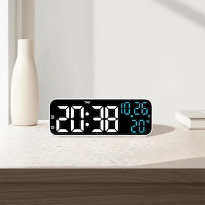 Peut inclure: Un réveil numérique rectangulaire à cadre blanc affiche l'heure, la date et la température. L'heure est affichée en grands chiffres blancs, tandis que la date et la température sont en petits chiffres bleus. La montre est sur une surface claire.