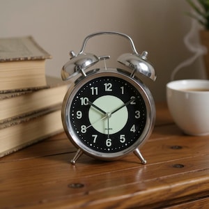 May include: A silver-colored alarm clock with a black face and white numbers. The clock has a round face with a glowing center and two bells on top. It sits on a wooden surface.