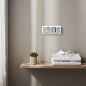 Puede incluir: Un reloj digital blanco que muestra las 8:13, 29,4 grados Celsius y 69% de humedad, montado en una pared marrón claro. Debajo, una estantería de madera con una pequeña planta en maceta, gafas y camisas blancas dobladas.