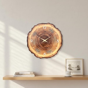 May include: A round wooden wall clock with a natural tree-trunk design. The clock face features a light brown color with darker brown concentric rings, white numerals, and gold-colored hands. The clock is mounted on a white wall.