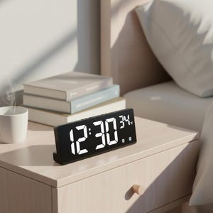 May include: A black digital alarm clock displaying 12:30 and 34 degrees Fahrenheit. The clock sits on a light wood nightstand next to a bed with a white pillow. A stack of books and a white mug are also on the nightstand.
