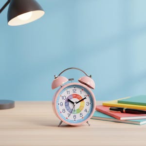 May include: A pink retro alarm clock with a rainbow-coloured inner dial and a light blue outer dial. The clock has a silver handle and two pink bells on top. The clock is on a wooden surface with notebooks and a pen.