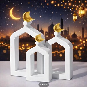 Puede incluir: Tres objetos decorativos blancos en forma de arco con detalles de luna creciente dorada. El fondo presenta una escena nocturna con una luna creciente, estrellas y siluetas de edificios, sugiriendo un tema cultural o religioso.