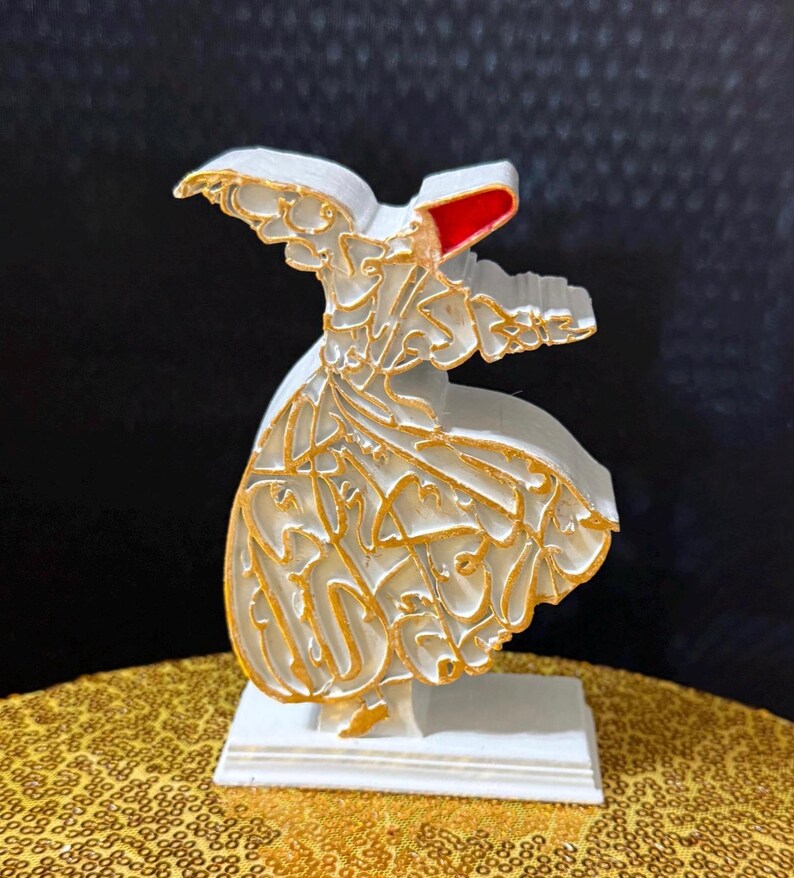 Puede incluir: Una figura blanca de un derviche giratorio con detalles dorados y un tocado rojo. La figura est&aacute; montada sobre una base blanca y presenta intrincada caligraf&iacute;a dorada. La figura se exhibe sobre una superficie dorada con lentejuelas.