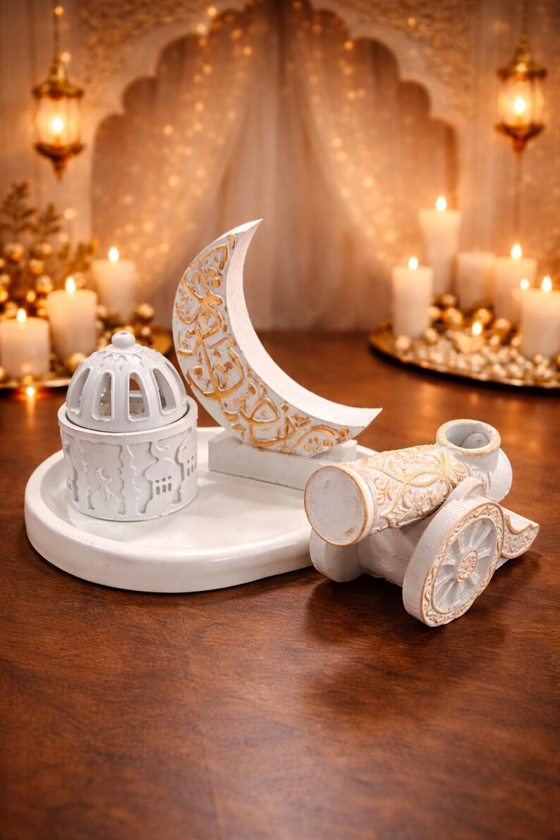 Puede incluir: Elementos decorativos blancos y dorados: una luna creciente, un ca&ntilde;&oacute;n y un recipiente con tapa sobre una bandeja blanca. La luna y el ca&ntilde;&oacute;n tienen detalles dorados. El fondo presenta velas y linternas.