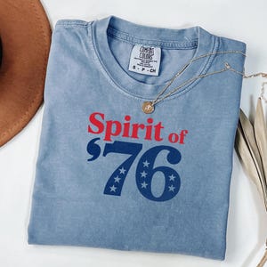 May include: A blue t-shirt with the text "Spirit of '76" in red and blue. The number 76 has stars in place of the zeros. A gold necklace with a pendant is on the shirt.