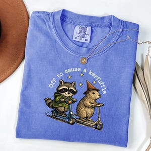 Könnte beinhalten: Blaues T-Shirt mit einem Aufdruck von einem Waschbären und einem Murmeltier auf Rollern, mit dem Text "Off to cause a kerfuffle". Der Waschbär trägt eine grüne Jacke und das Murmeltier einen Zaubererhut. Eine goldene Halskette liegt auf dem T-Shirt.