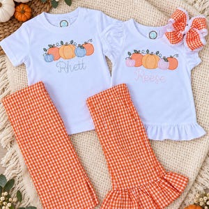 May include: Two white short-sleeved shirts with embroidered pumpkin designs and the names Rhett and Reese, paired with orange and white gingham pants. One shirt has a ruffled hem and a matching orange and white gingham hair bow is included.