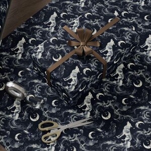 Op de afbeelding: Donkerblauw cadeaupapier met een herhalend patroon van witte figuren, manen en wolken. Een ingepakt cadeau met een bruin lint ligt op het papier, samen met een rol van hetzelfde patroon. Een gouden schaar en een zilveren plakbanddispenser liggen ook op de tafel.