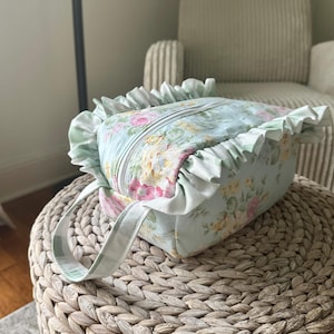 May include: A floral print cosmetic bag with a light blue background and a zipper closure. The bag features a ruffle trim and matching handles. The floral pattern includes pink, yellow, and white flowers.