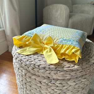 May include: A square pillow with a yellow bow and ruffle trim. The pillow has a floral and polka dot pattern in yellow and blue. The pillow is resting on a woven, beige ottoman.