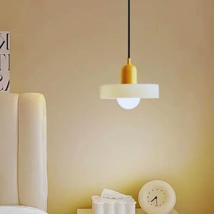 May include: A modern pendant light fixture with a yellow top and a white, disc-shaped shade. The light hangs from a black cord and illuminates a soft, warm glow. The fixture is a stylish addition to any interior space.