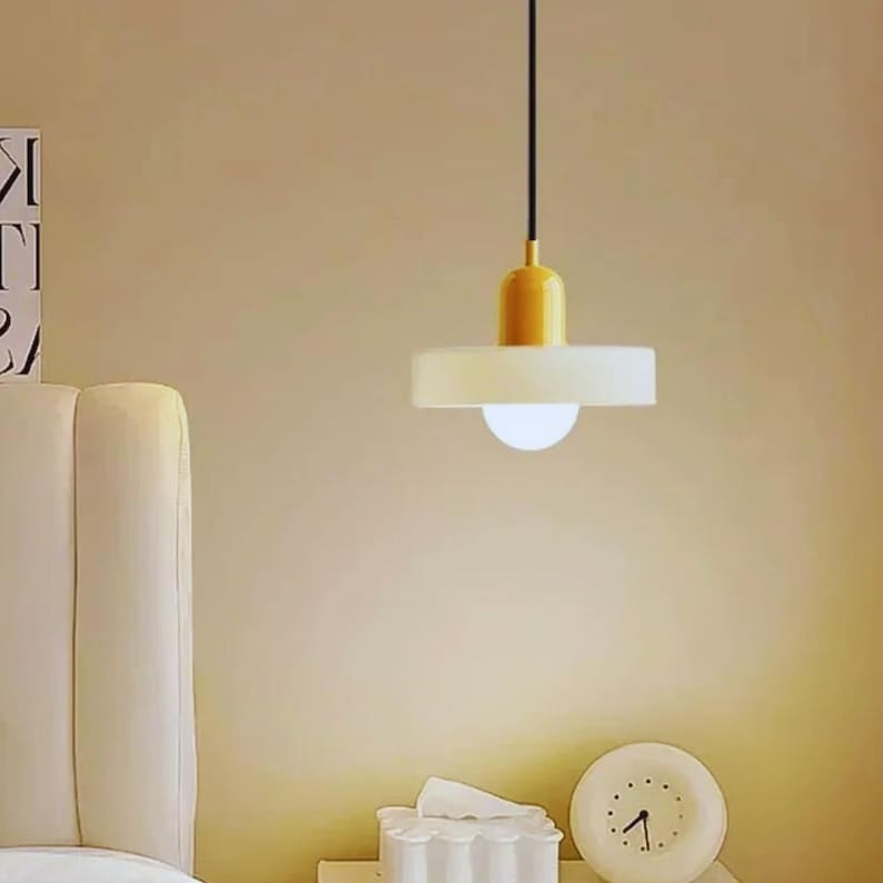 May include: A modern pendant light fixture with a yellow top and a white, disc-shaped shade. The light hangs from a black cord and illuminates a soft, warm glow. The fixture is a stylish addition to any interior space.