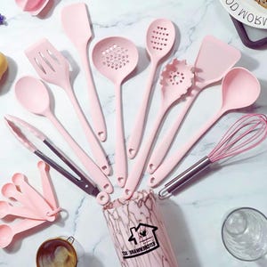 May include: A selection of pink kitchen utensils, such as spatulas, spoons, and a whisk, displayed on a white marble surface. The set includes various tools for cooking and baking, ideal for any kitchen.