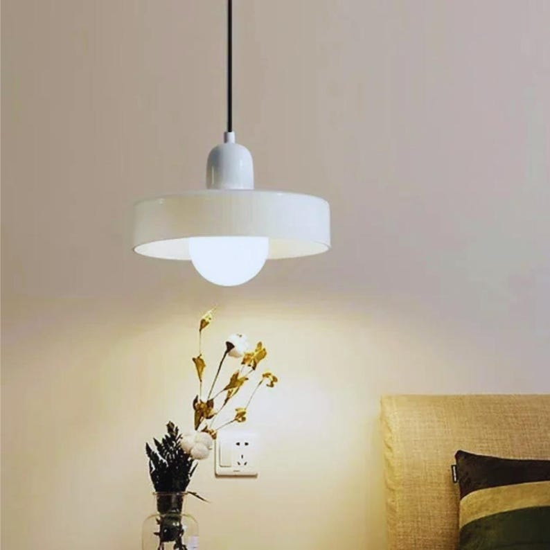 May include: A white, modern pendant light fixture with a circular shade and a spherical bulb. The light hangs from a black cord. The image also shows a vase of flowers and a neutral-toned wall.