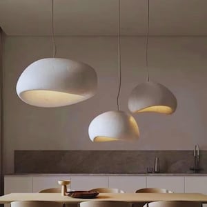 May include: Three white, organically shaped pendant lights hang from the ceiling. The lights have a textured surface and a warm glow from the open bottom. The lights are suspended above a dining table in a kitchen setting.