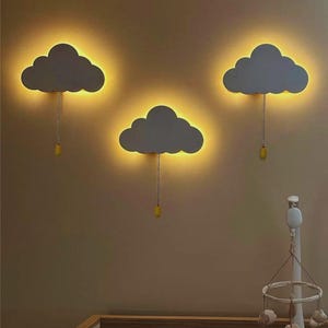Könnte beinhalten: Drei graue Wolken-Wandleuchten mit warmgelbem Schein. Jede Leuchte hat eine Kette mit einem gelben Zug. Die Leuchten sind an einer beigefarbenen Wand montiert und erzeugen einen weichen, umgebenden Lichteffekt. Teil einer Kinderzimmerdekoration.