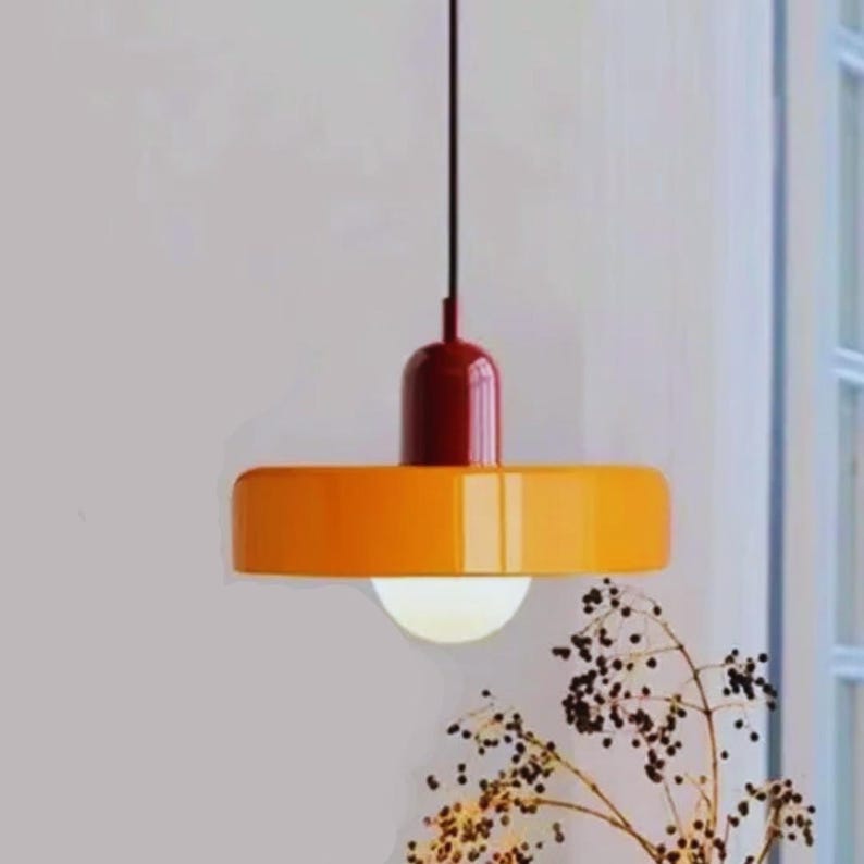 May include: A modern pendant light fixture with a circular, orange shade and a dark red top. The light hangs from a black cord and has a white, spherical bulb. The fixture is a stylish addition to any home decor.