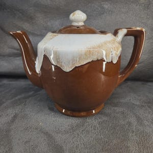 May include: A ceramic teapot featuring a glossy brown base and a white drip glaze pattern around the top and lid. The teapot has a curved spout, a sturdy handle, and a small knob on the lid, showcasing a two-tone color scheme throughout.
