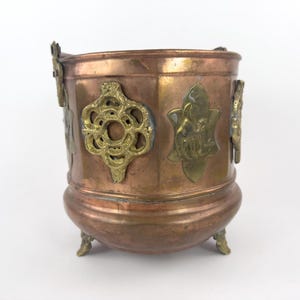 May include: A copper-colored metal planter with ornate brass accents. The planter has a rounded base and three small feet. Decorative brass elements include floral and geometric designs. The planter is likely vintage or antique.