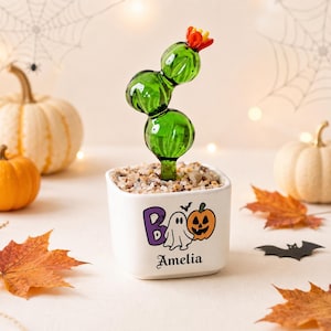 Op de afbeelding: Een witte vierkante keramische plantenbak gevuld met kleine steentjes en een groene glazen cactussculptuur met een rode bloem. De plantenbak is voorzien van een afbeelding met de tekst BOO, een wit spookje, een pompoen en de naam Amelia in zwart.