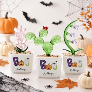 May include: Three white square ceramic planters, each containing a unique glass plant sculpture. The planters feature the text "Boo" with a pumpkin and ghost graphic, and the name "Kathryn" printed below. The glass plants include a pink flower, a green cactus, and a curved green stem with purple flowers.