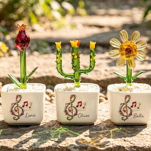 May include: Three glass plant figurines in white square pots. Each pot features a floral treble clef and musical notes with the names Luna, Oliver, and Bella. The plants include a red flower, a green cactus, and a yellow daisy, all set in white gravel.