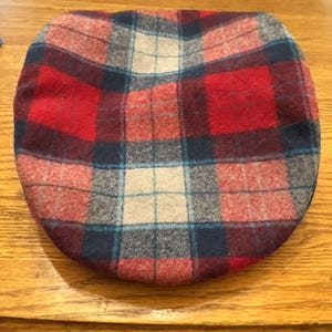 May include: A flat cap with a plaid pattern in red, navy blue, light blue, and beige. The cap is made of a woven fabric and is likely wool or a wool blend. The cap is a classic style.