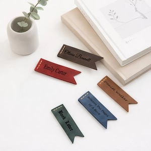 May include: Several leather bookmarks in shades of red, brown, and blue, each with a name or phrase. The bookmarks include names like "Emily Carter" and phrases like "Book Lover". The bookmarks are made of leather.