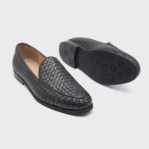 Puede incluir: Un par de mocasines negros con una textura superior de cuero tejido. Un zapato está colocado en posición vertical para mostrar el perfil superior, mientras que el otro está boca abajo para revelar la suela negra con patrones de tracción circulares.