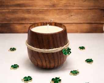 May include: A wooden candle holder with a lit white candle, wrapped with twine and a green shamrock charm. Several green shamrock charms are scattered around the candle holder. The background is a wooden wall.