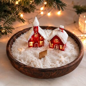 以下が含まれることがあります： 人工の白い雪で満たされた暗い木製の丸いボウル。中には、窓が光り、屋根に雪が積もった2つの赤いミニチュアハウスが入っています。片方の家には星の切り抜きがある尖塔があり、もう片方の家には煙突があります。