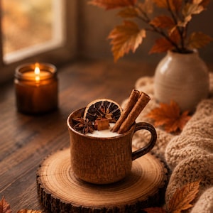 May include: A brown ceramic mug candle featuring a wooden wick, topped with a dried orange slice, star anise, and two cinnamon sticks. The candle sits on a circular wood slice coaster, surrounded by scattered autumn leaves and a knit fabric.