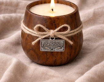 May include: A lit candle in a wooden barrel-shaped holder. The brown holder is wrapped with twine and a small metal charm that reads "Friendship". The candle sits on a beige cloth.